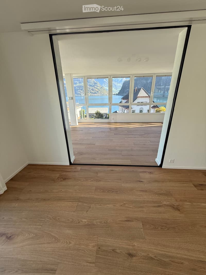 Empty room with wooden flooring and large sliding glass doors leading to a balcony with scenic mountain and lake views.