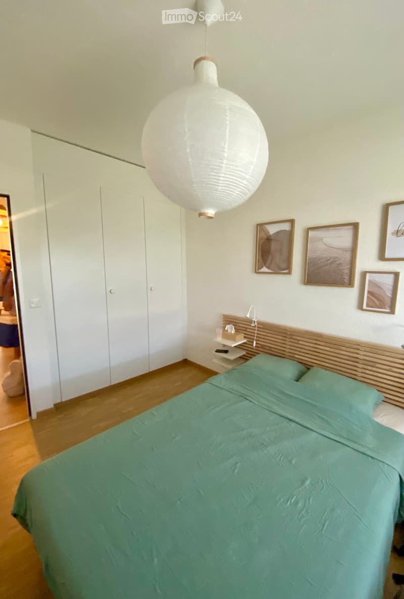 Modern bedroom with a green duvet-covered bed, wooden nightstand, pendant light, and framed pictures on the wall.
