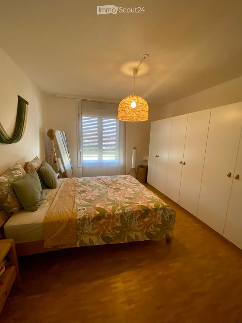Bright bedroom with wooden flooring, a large bed with a floral duvet and pillows, a wardrobe, a mirror, and a hanging lamp with a woven shade.