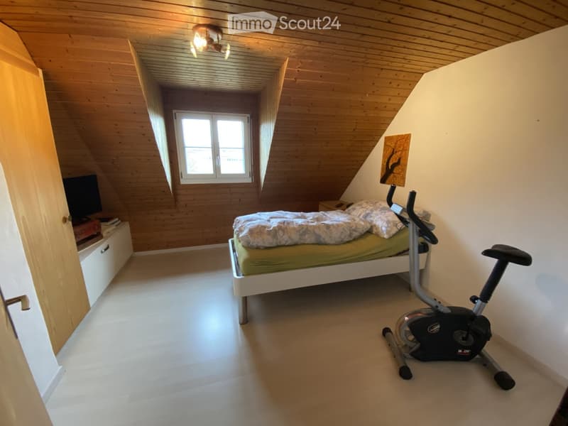 Cozy attic bedroom with wooden slanted ceilings, a single bed, exercise bike, nightstand, and a TV screen on a stand.