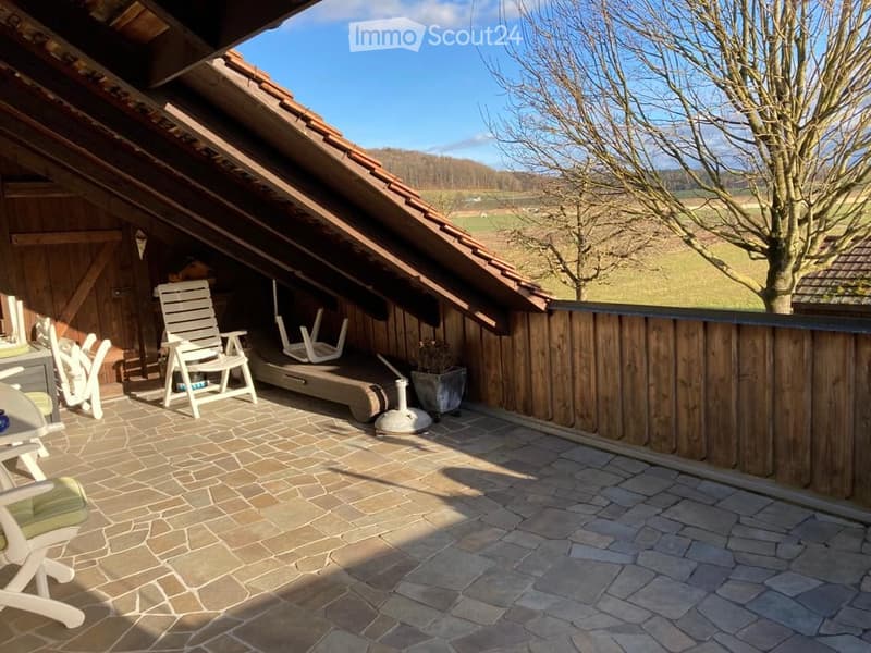 Wooden balcony with stone tiles, white chairs, small table, and potted plant, overlooking a rural landscape.