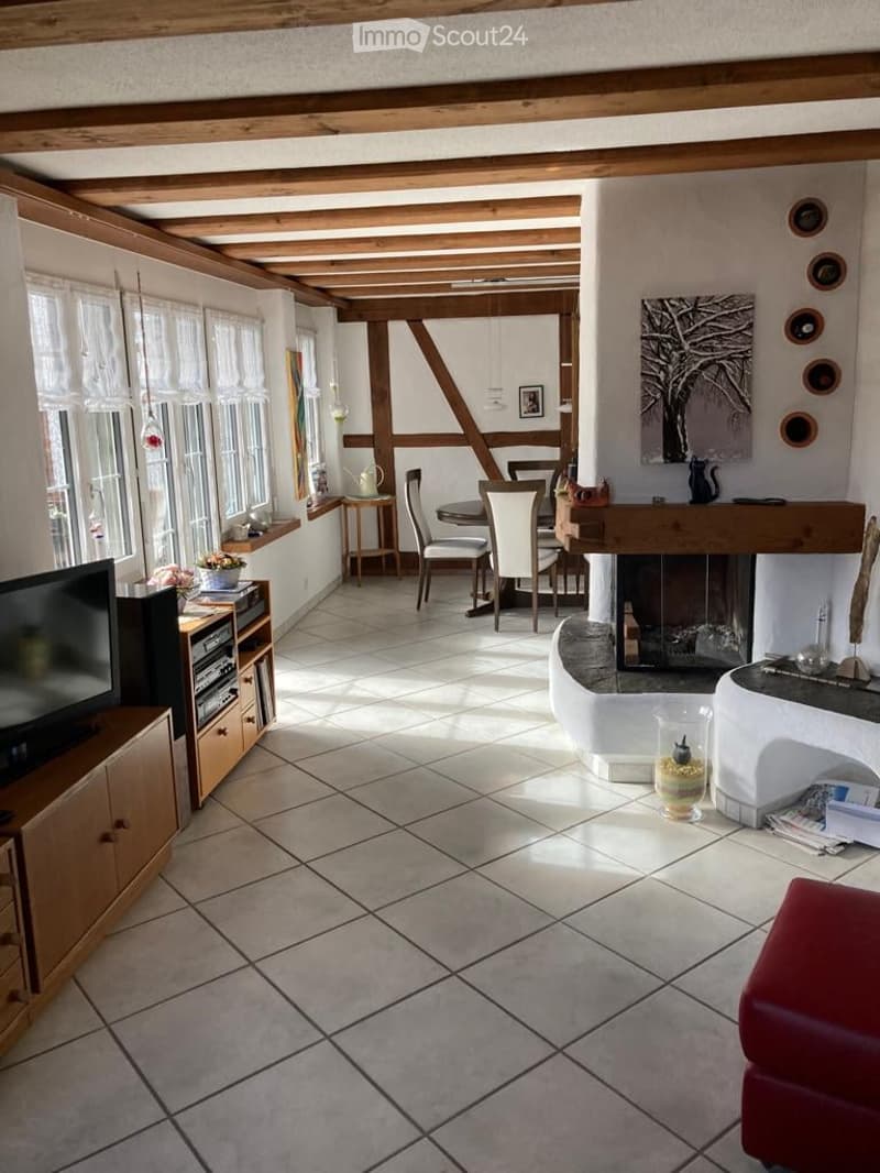 A cozy living room with exposed wooden beams, a fireplace, a television, a dining area with chairs and a table, and a small seating area with a red chair and a vase on a table.