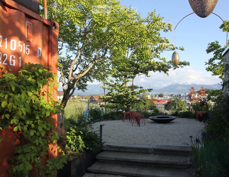 Garden area with gravel pathway, steps, red stools, fire bowl, and hanging decorative light, surrounded by greenery and a view of mountains.