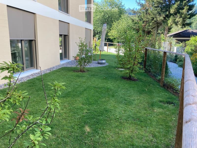 Well-maintained garden with green grass, small trees, a wooden fence, and a modern house with large windows and a sliding door.