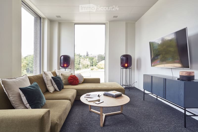 Modern living room with large windows, grey carpet, beige sectional sofa, wooden coffee table, floor lamps, and a flat-screen TV mounted on the wall.