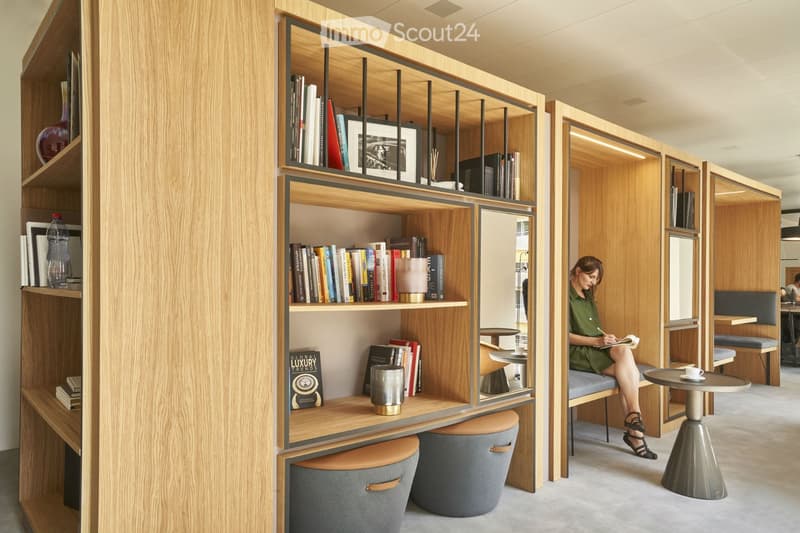 Modern, minimalist room with wooden shelves filled with books and decorative items, a small round table with a cup, and a comfortable bench with a person writing.