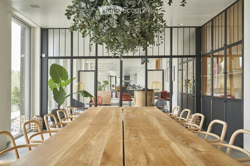 A modern, well-lit room with large glass partitions, a long wooden table surrounded by chairs, and a hanging plant. The space features contemporary decor, including a mirror and a screen in the background.
