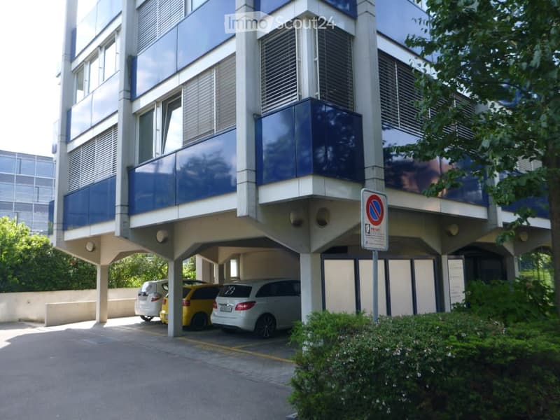 modern building, blue facade, glass windows, covered parking area, yellow and white cars parked, bushes and trees
