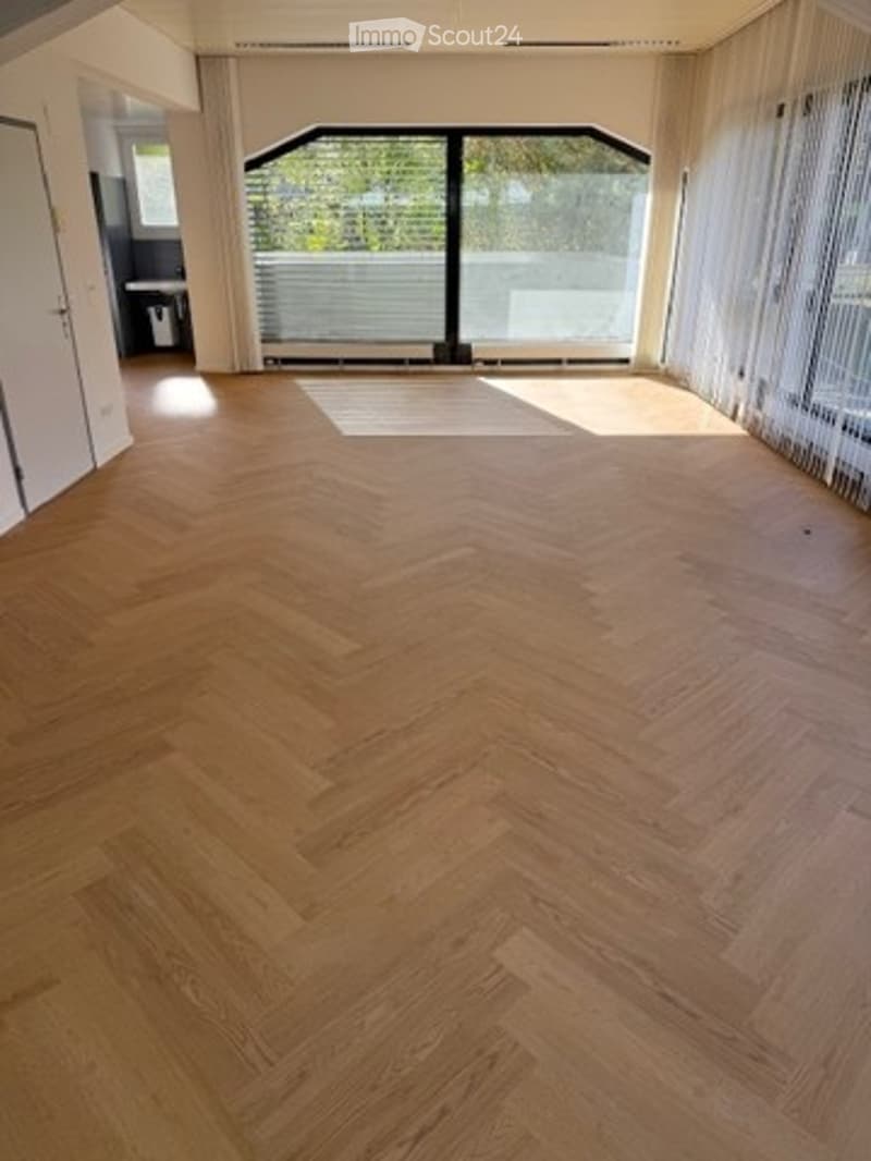 Empty room with herringbone-patterned wooden flooring, large windows with white blinds, and white walls.
