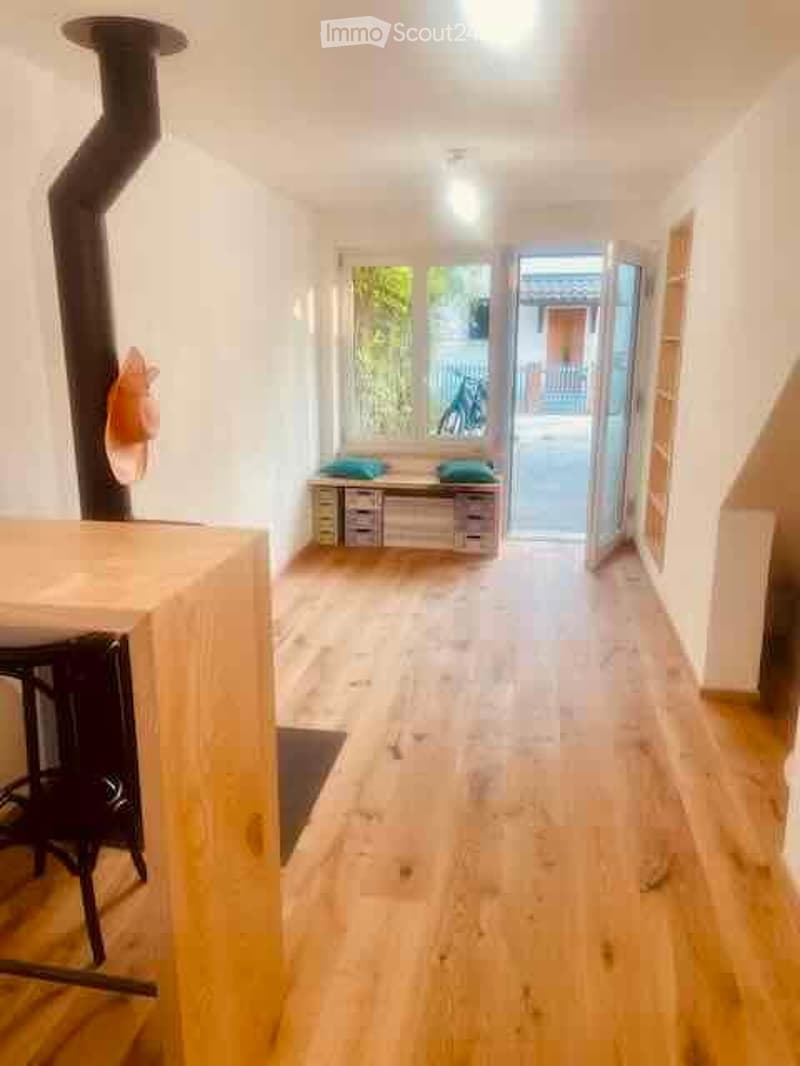Bright hallway with wooden flooring, white walls, and a bench with cushions near a large window. A black chair is visible on the left side.