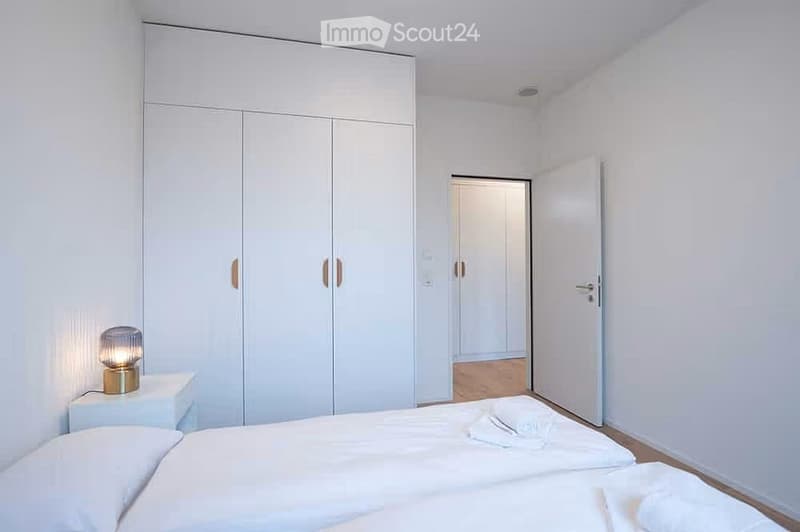 Modern, minimalist bedroom with a large bed, white nightstand, lamp, and built-in white wardrobe.