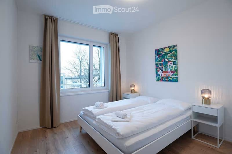 Bright bedroom with a double bed, two nightstands with lamps, and large windows with curtains. The room features a colorful painting on the wall and a wooden floor.