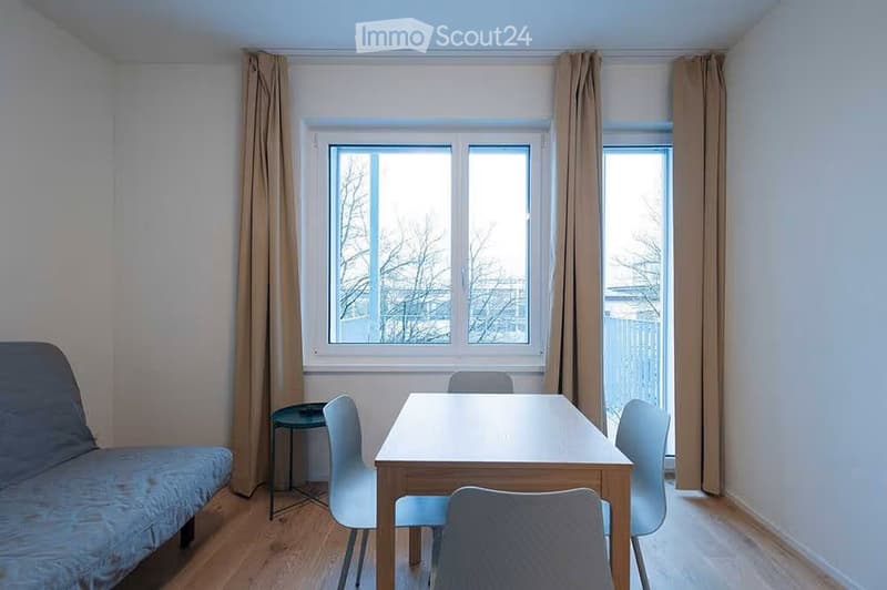 A small, furnished room with a sofa, a table, and chairs, featuring large windows with curtains and wooden flooring.