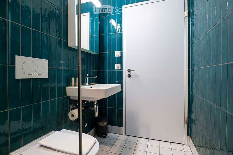 Modern bathroom with dark green tiled walls, white tiled floor, toilet, sink with chrome faucet, mirror, and a white door.