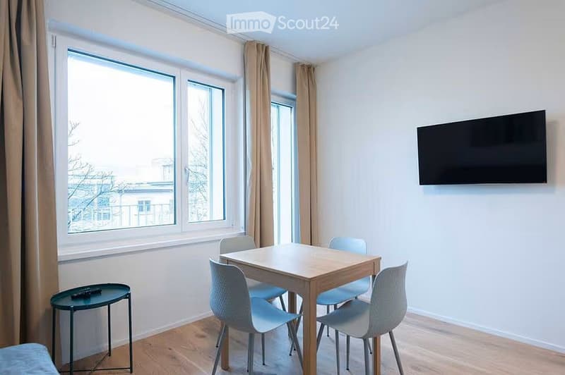 Bright room with large windows, a wooden dining table with four chairs, a small round stool, and a wall-mounted TV.