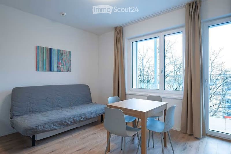 A small, furnished room with a sofa, a small table with chairs, and large windows with curtains. The room has a modern, minimalist design with light-colored walls and wooden flooring.