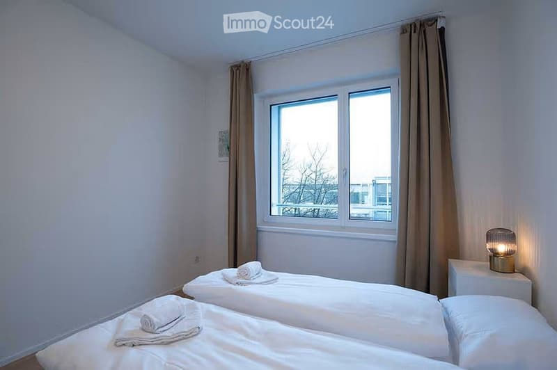 Bright bedroom with two single beds, white linens, and a nightstand with a lamp. The room has large windows with curtains and a view of the outside.