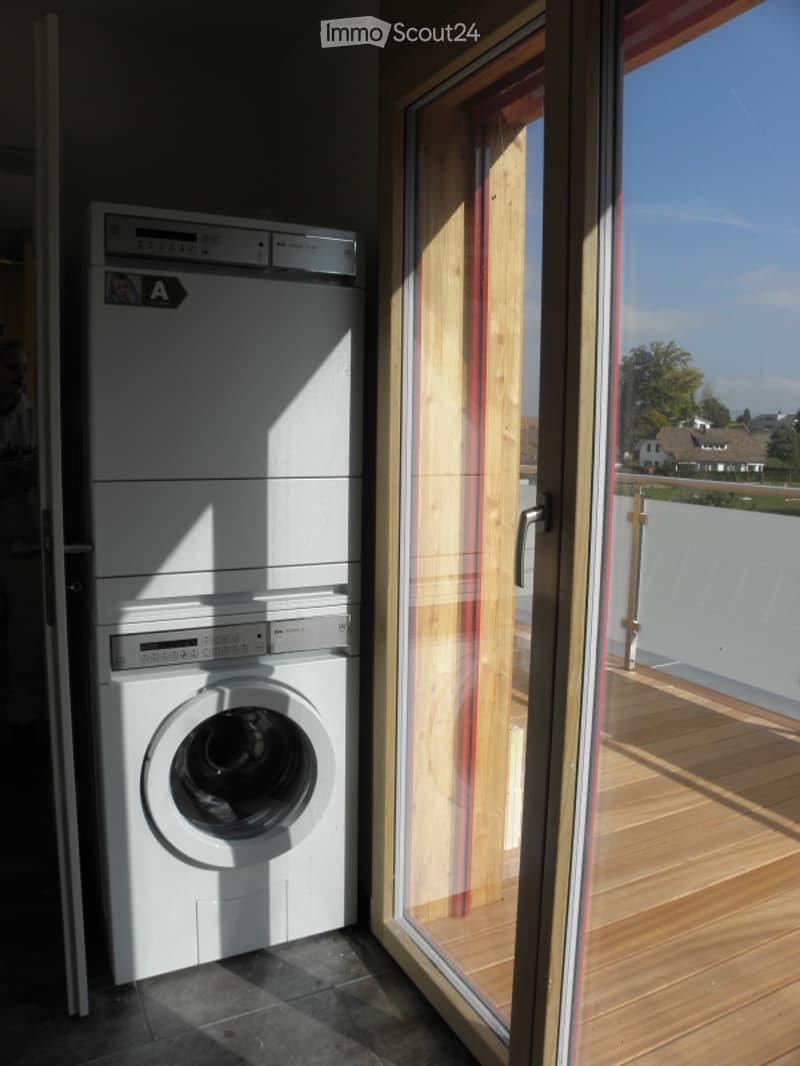 Modern bathroom with a washing machine and dishwasher stacked vertically, large window with view, wooden floor, and tiled walls.