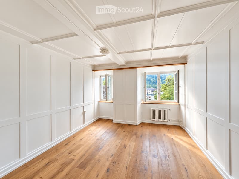 An empty room with white walls, wooden flooring, and large windows offering a view of greenery. The room features a radiator and a decorative wooden ceiling.