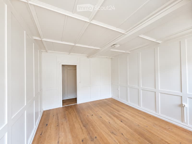 Empty room with white paneled walls, wooden floor, and a white ceiling with decorative molding. No furniture or equipment present.