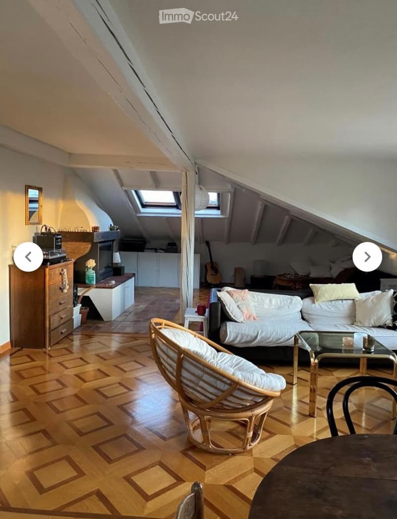 Spacious attic room with wooden flooring, slanted ceilings, and large windows. Features a sofa, wicker chair, coffee table, guitar, and a small kitchen area with cabinets and a sink.