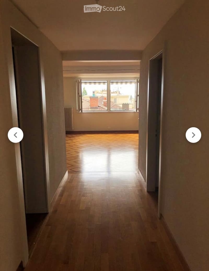 A long, narrow hallway with wooden flooring and beige walls, leading to a room with large windows and a view of rooftops.