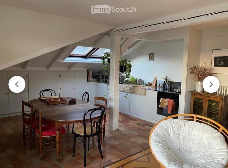 A cozy attic space with a dining area and a small kitchen. The dining area features a wooden table with chairs, and the kitchen has white cabinets, a sink, and an oven. The room has exposed wooden beams and skylights, creating a bright and airy atmosphere