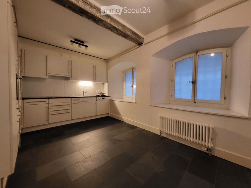 Empty kitchen with white cabinets, dark tiled floor, and large windows. No furniture or appliances are present.