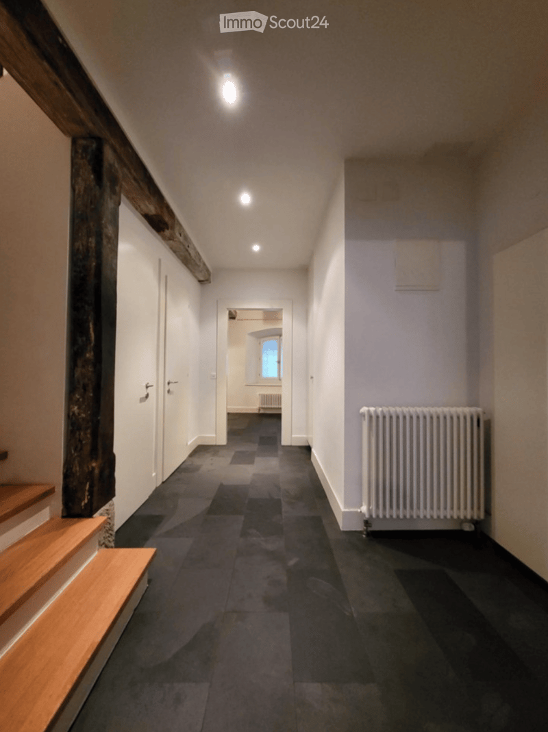 A narrow hallway with wooden stairs on the left, dark flooring, white walls, and a radiator on the right side. The hallway leads to an open doorway at the end.