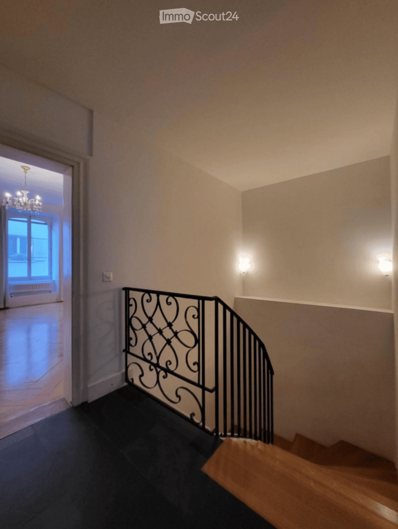 Spacious hallway with wooden staircase, black wrought-iron railing, and warm lighting. The hallway features beige walls and a dark carpeted floor, leading to an open doorway with a chandelier visible in the adjacent room.