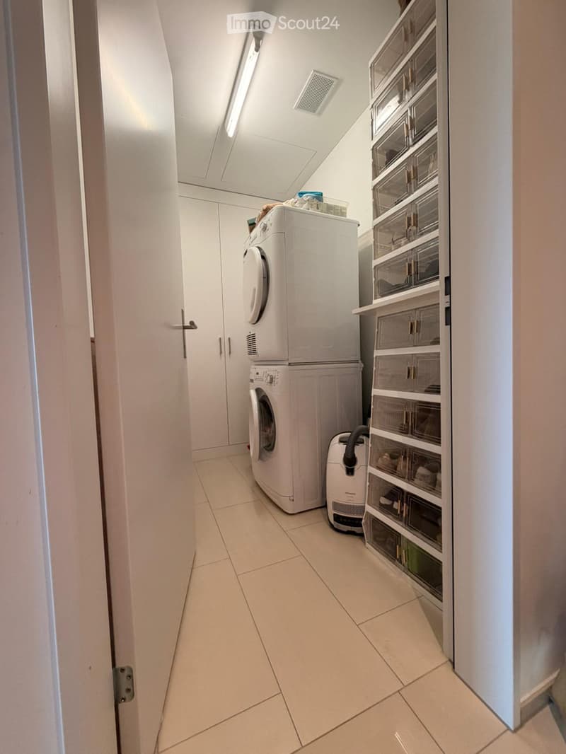 A narrow hallway with tiled flooring, white walls, and a door on the left. A washing machine and dryer are positioned against the right wall, accompanied by a vacuum cleaner and built-in storage cabinets.