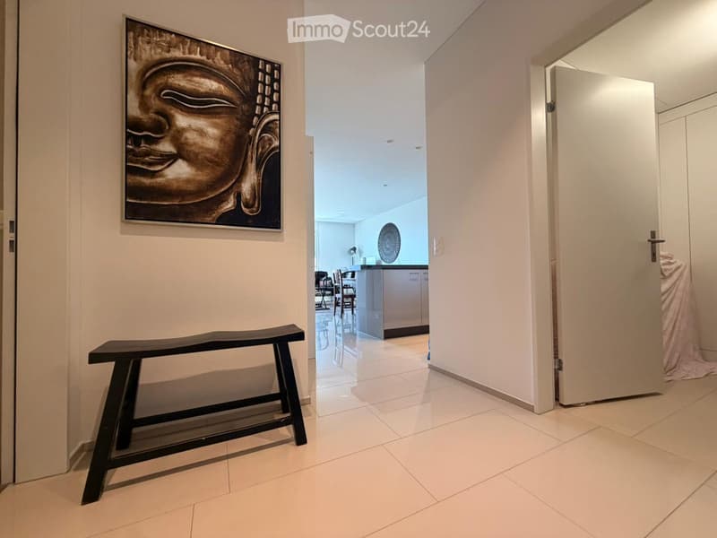 A modern hallway with white tiled flooring, a black bench, and a large Buddha head artwork on the wall. The hallway leads to a kitchen area with white cabinets and a dining table with chairs.