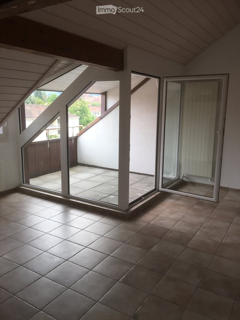 Empty room with tiled floor and large windows opening to a balcony area. The room has a slanted ceiling typical of an attic space.