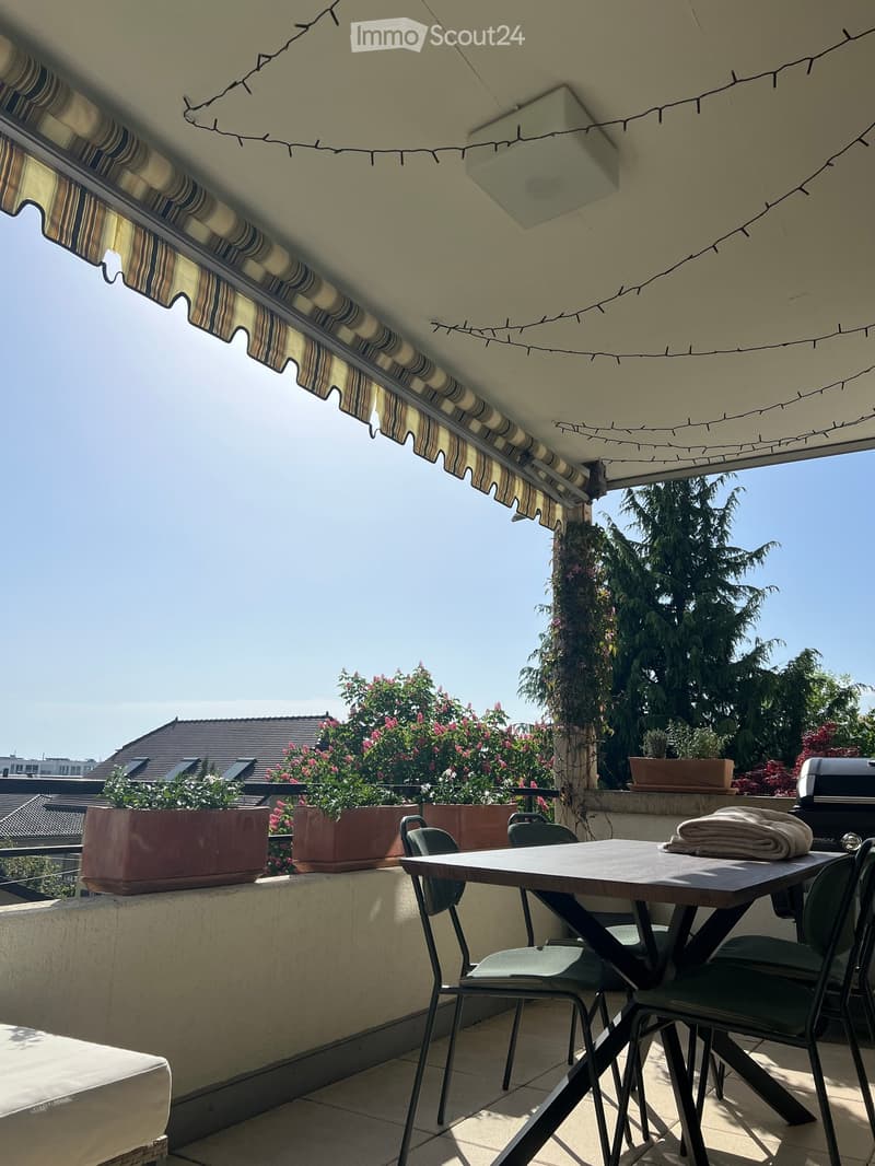 Balcony with a table, chairs, potted plants, and a grill, featuring a striped awning and string lights.