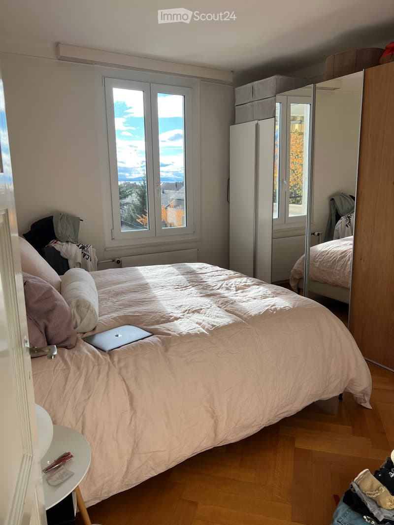 A well-lit bedroom with a large bed, laptop on the bed, a mirror, wardrobe, and a small table with items on it.