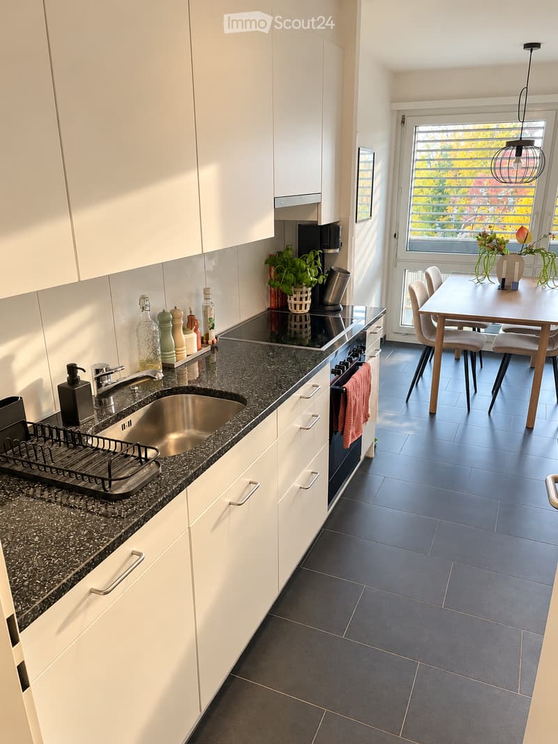 Modern kitchen with white cabinets, dark granite countertops, stainless steel sink, and built-in appliances. A small dining area with a table and chairs is visible through an open doorway.