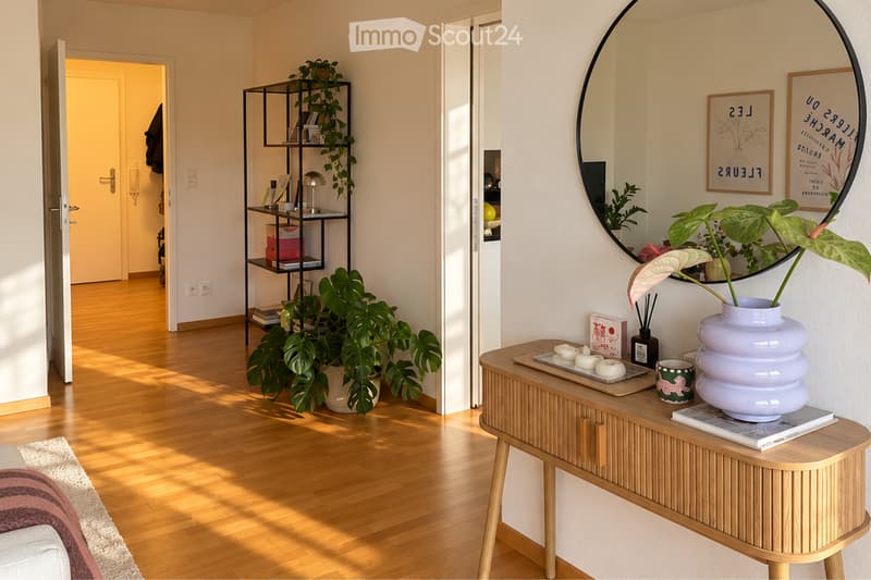 A modern, well-lit hallway with wooden flooring, a round mirror on the wall, a small table with a vase and candles, a potted plant, and a shelf with books and decorative items.