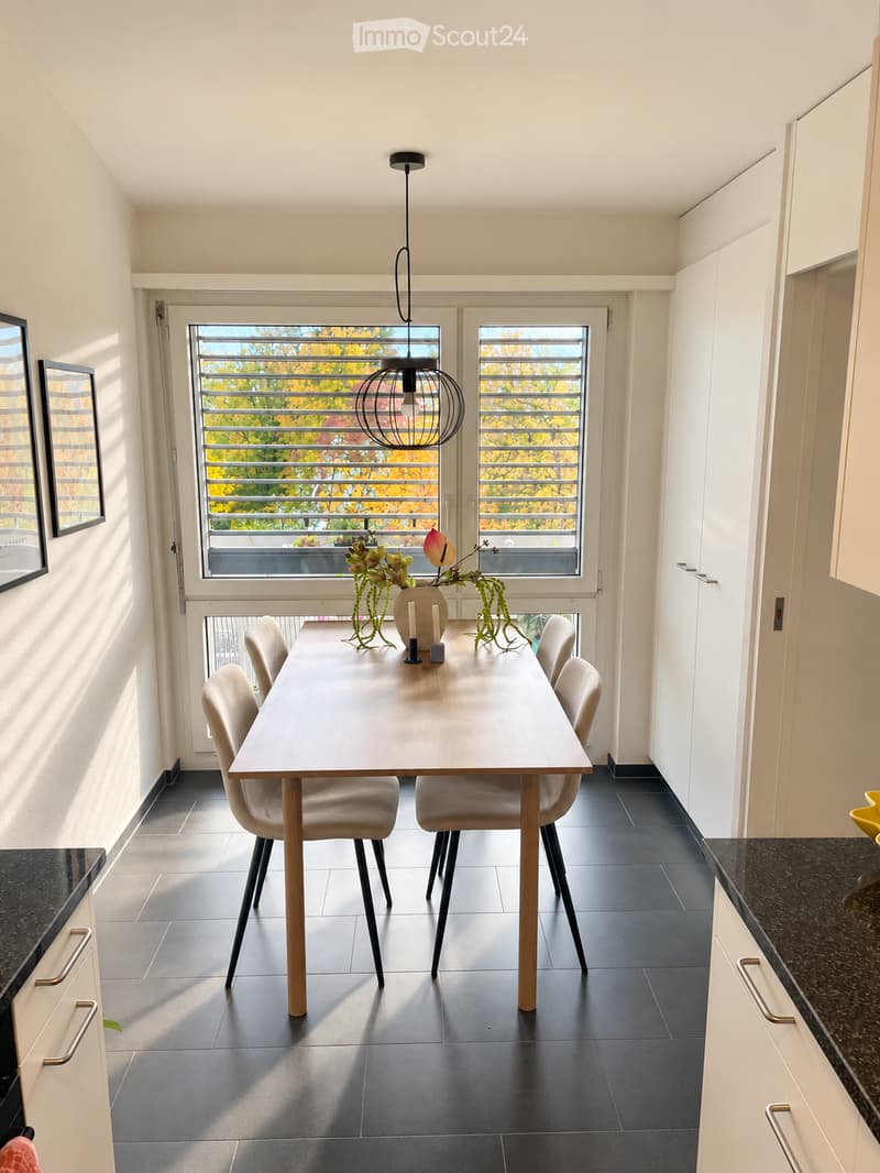 Modern dining area with a light wood dining table, six chairs, a hanging pendant light, and a vase with flowers on the table. The room features dark tiled flooring and white cabinetry.