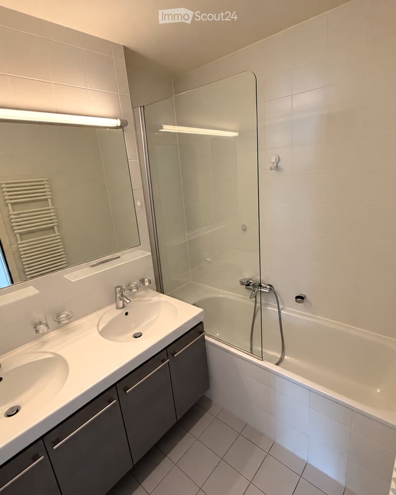 Modern bathroom with a double sink, bathtub, large mirror, and a radiator. The space features tiled walls and a glass-enclosed bathtub area.