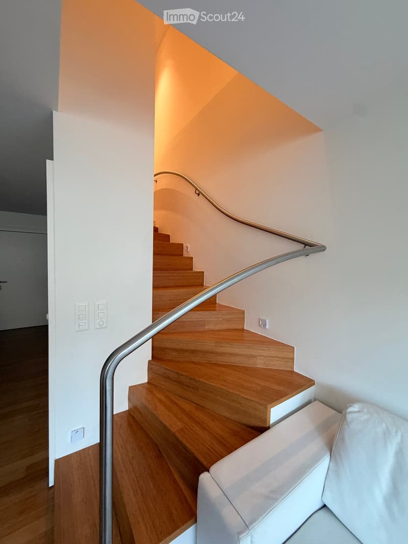 Modern staircase with wooden steps and a sleek metal handrail, leading to an adjacent room with a visible sofa.