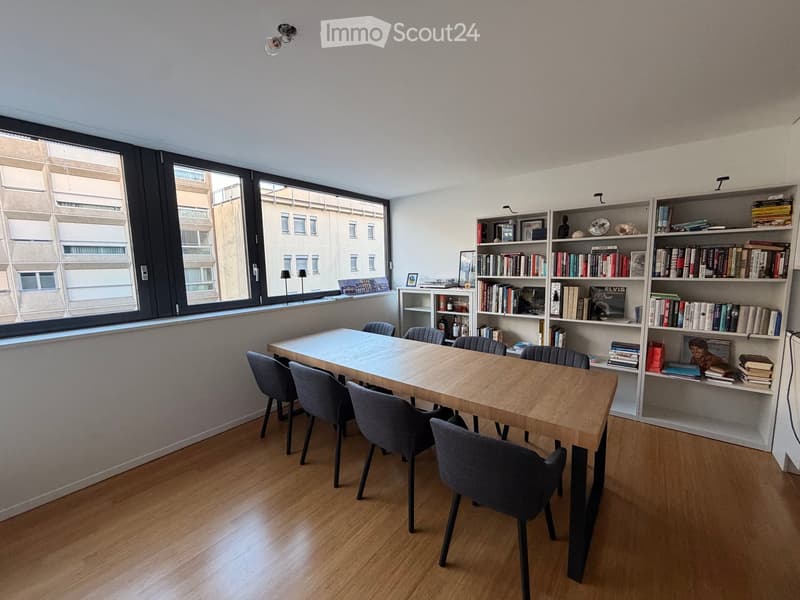Bright room with wooden floor, large window, wooden table with six chairs, bookshelf filled with books and decorative items, and two table lamps near the window.