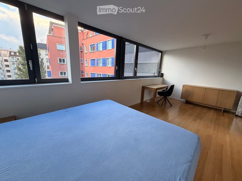 A modern bedroom with a blue bedspread, wooden floor, large windows, a small desk with a chair, and a wooden cabinet.