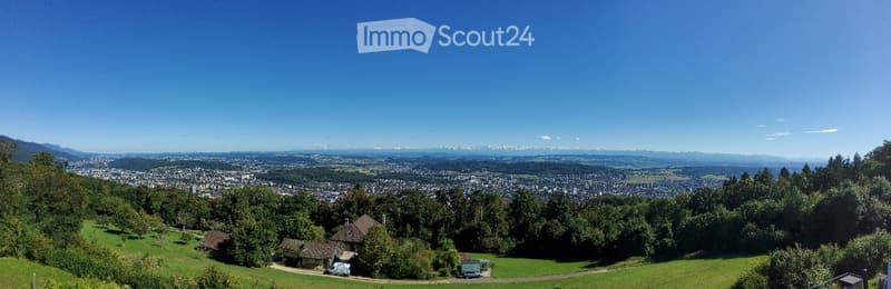 Panoramic view of a town and surrounding landscape from a hilltop, with houses, trees, and distant mountains visible.