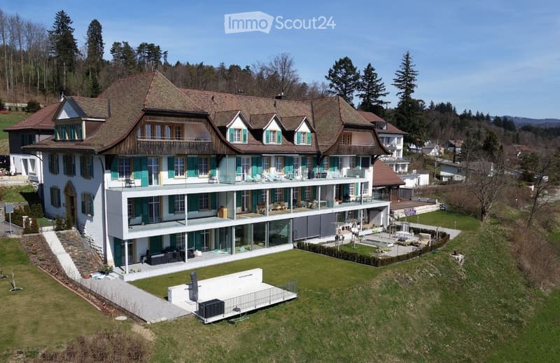 A large, two-story house with a modern extension featuring large glass windows and a spacious balcony. The traditional section has a pitched roof and green shutters. The property includes a well-maintained lawn, patio area, and surrounding trees.