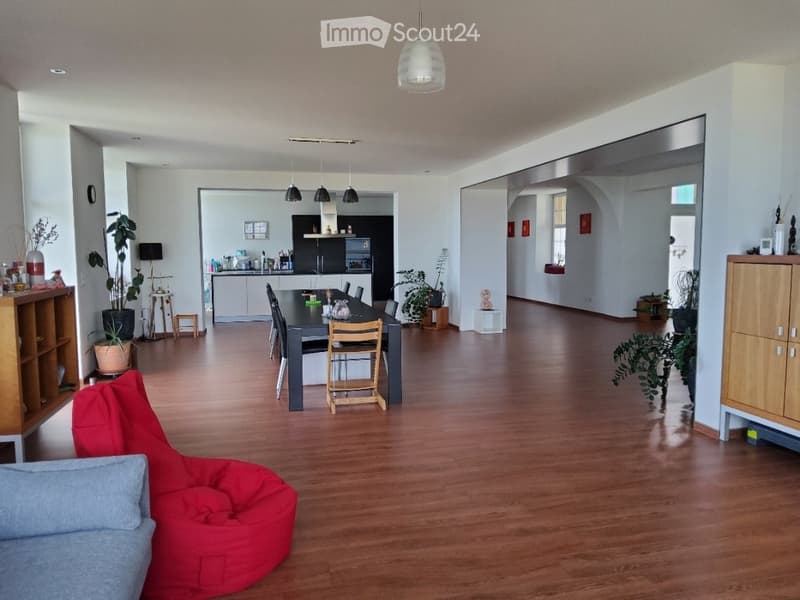 Open-plan living and dining area with wooden flooring, a red bean bag chair, a dining table with chairs, and a modern kitchen in the background.