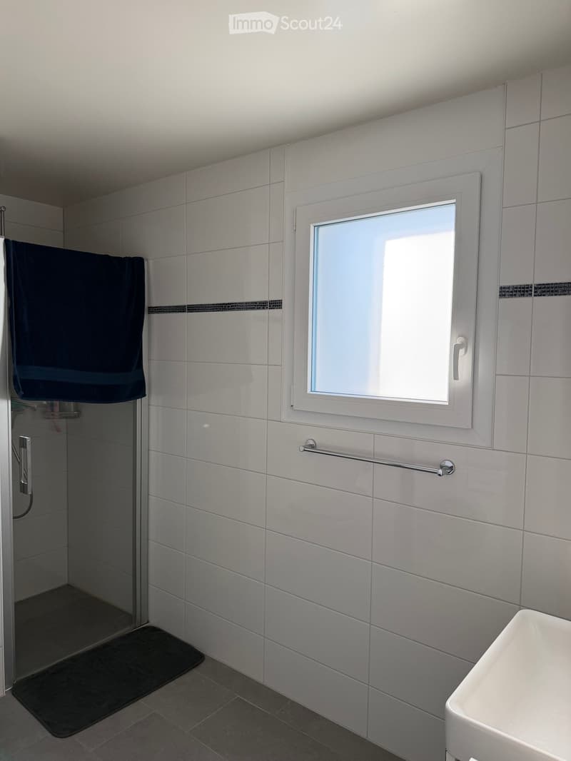Modern bathroom with white tiles, a window, a sink, a towel, and a shower area with a glass door.
