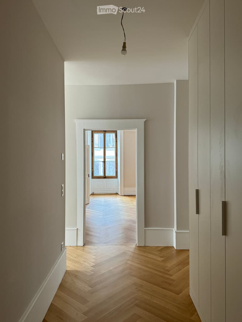 A modern hallway with light wooden herringbone flooring, white walls, and a single hanging light fixture. An open doorway leads to another room with similar flooring and white walls.