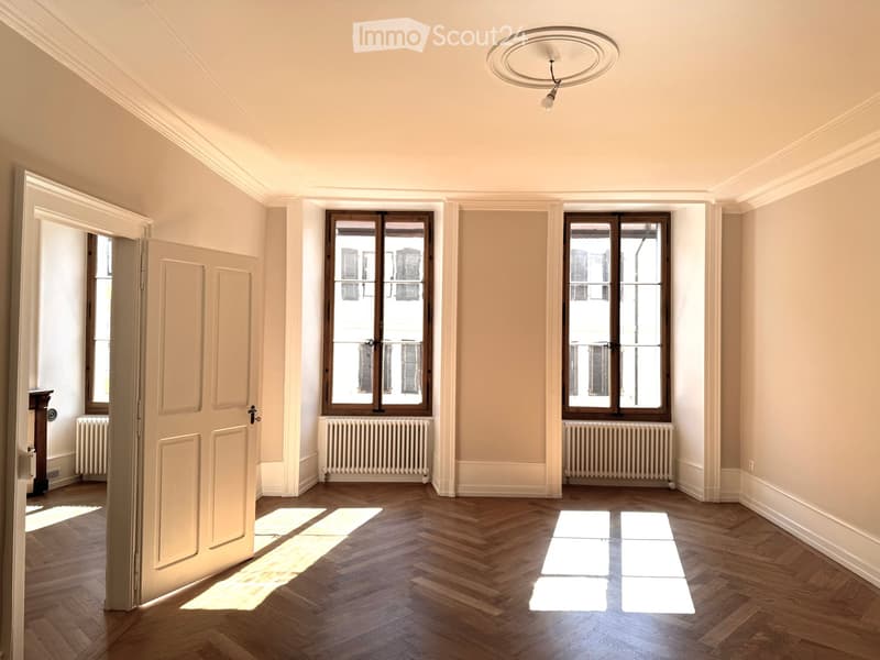 Empty room with wooden flooring, large windows, radiators, and a ceiling light fixture.