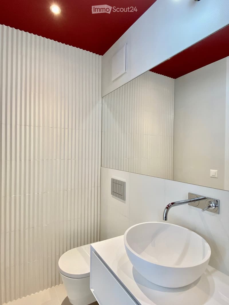 Modern bathroom with a white toilet, a sleek white sink bowl, and a chrome faucet mounted on a white wall. The walls are white with vertical ribbed patterns, and the ceiling has a red and white design with a skylight window.