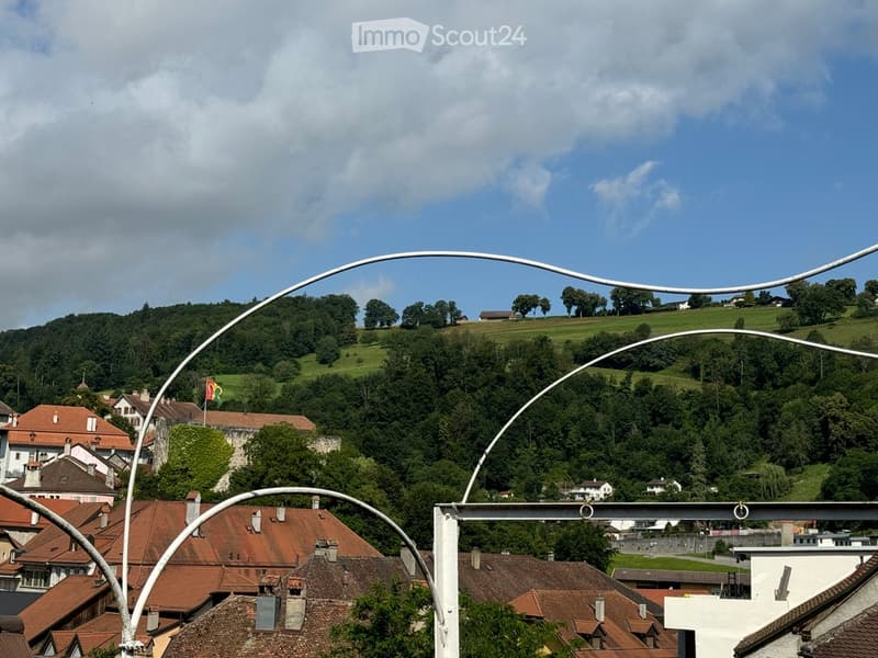 View of a town with red-tiled roofs, surrounded by green hills and trees under a partly cloudy sky.